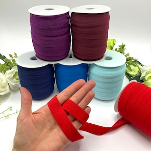 May include: Spools of elastic ribbon in various colors: purple, burgundy, navy blue, turquoise, and light blue. A hand holds a length of red elastic ribbon, showcasing its texture and color. The spools are on a white surface, with a red spool in the foreground.
