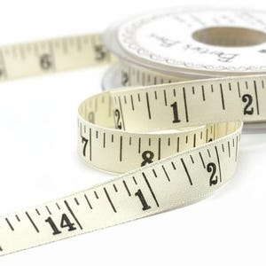 Pode incluir: Fita métrica de cor marfim com marcações pretas, desenrolada de um carretel de plástico transparente. A fita mostra medidas em polegadas, com números e traços. O carretel está no fundo e a fita em primeiro plano.