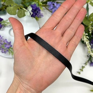 May include: A close-up of a hand holding a black satin ribbon. The ribbon is smooth and shiny, and it is about 1/2 inch wide.