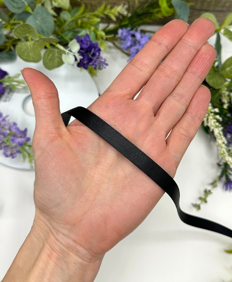 May include: A close-up of a hand holding a black satin ribbon. The ribbon is smooth and shiny, and it is about 1/2 inch wide.