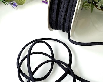 Puede incluir: Un carrete de cordón elástico negro, con una porción desenrollada y en bucle. El cordón es de color oscuro sólido y parece estar hecho de un material flexible similar a la tela. El fondo es blanco, con algo de vegetación y flores moradas.