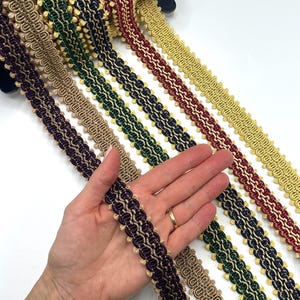 Puede incluir: Una selección de cintas decorativas de diferentes colores, incluyendo dorado, burdeos, verde y morado. La cinta presenta un diseño en bucle con un borde contrastante. Las cintas se muestran sobre una superficie blanca.
