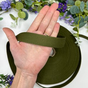 May include: Olive green canvas webbing ribbon held in a hand. The ribbon is rolled on a cardboard core. The background includes purple and green floral accents. This durable webbing is suitable for various crafting and sewing projects.
