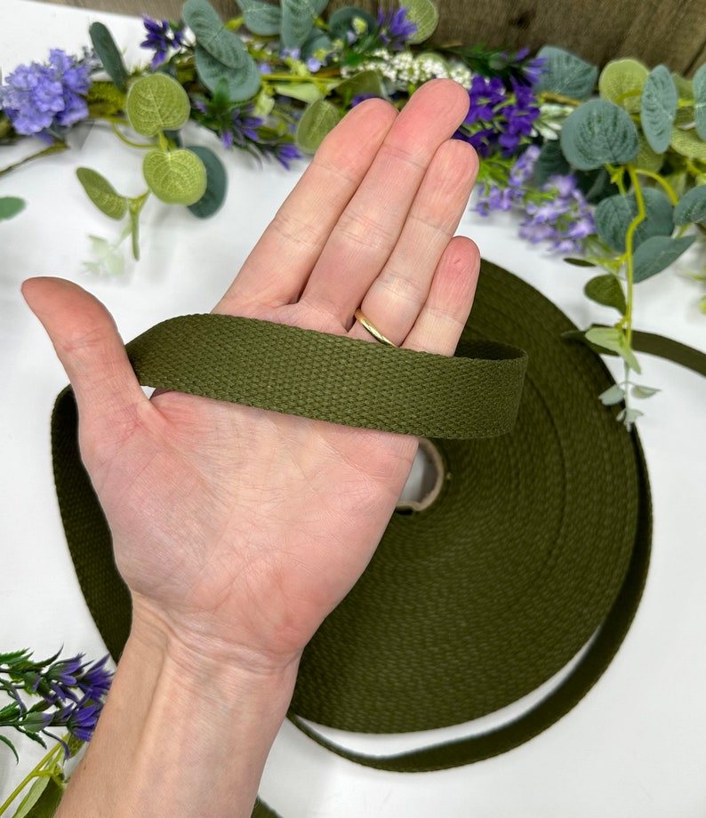 May include: Olive green canvas webbing ribbon held in a hand. The ribbon is rolled on a cardboard core. The background includes purple and green floral accents. This durable webbing is suitable for various crafting and sewing projects.