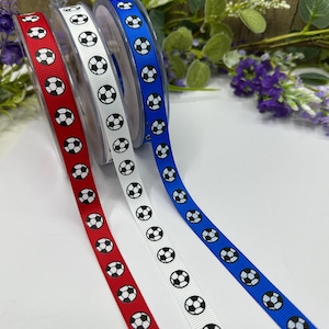 Pode incluir: Três rolos de fita em vermelho, branco e azul, cada um adornado com um padrão repetido de bolas de futebol em preto e branco. As fitas são exibidas em um fundo de vegetação e flores roxas.