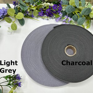 May include: Two spools of woven webbing in light grey and charcoal colors. The light grey spool is labeled "Light Grey" and the darker spool is labeled "Charcoal". The spools are displayed on a white surface with floral accents.