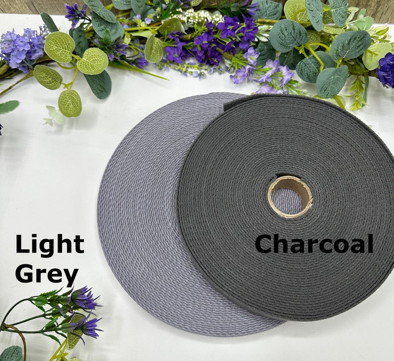 May include: Two spools of woven webbing in light grey and charcoal colors. The light grey spool is labeled "Light Grey" and the darker spool is labeled "Charcoal". The spools are displayed on a white surface with floral accents.