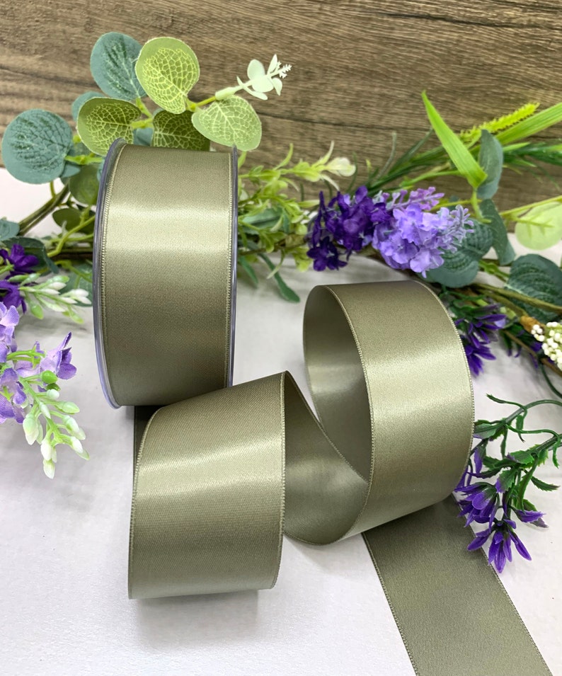 May include: A roll of sage green satin ribbon. The ribbon is displayed on a white surface with green foliage in the background.