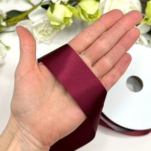 May include: A spool of burgundy satin ribbon. The ribbon is 1 inch wide and is shown wrapped around a hand.