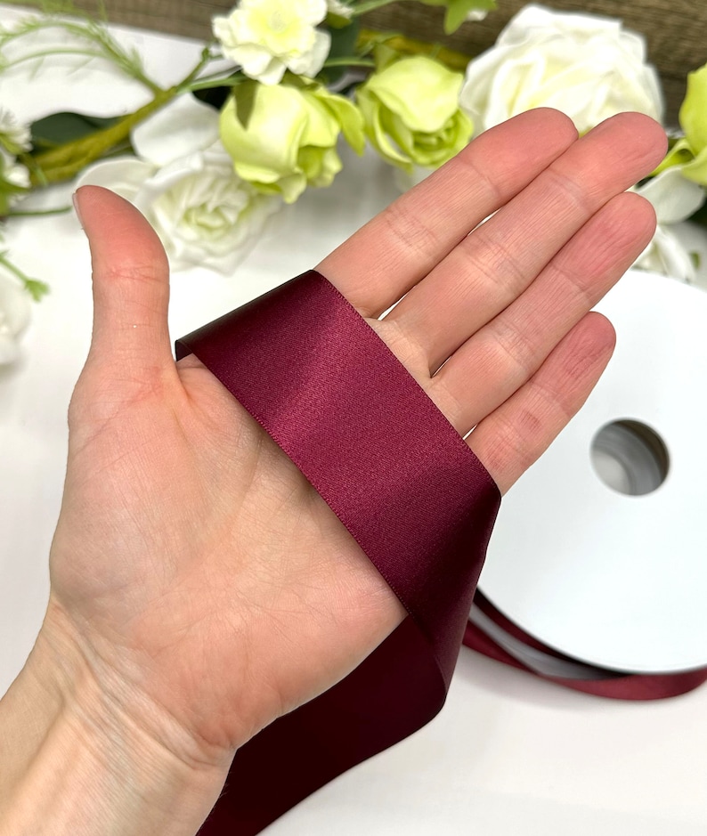 May include: A spool of burgundy satin ribbon. The ribbon is 1 inch wide and is shown wrapped around a hand.