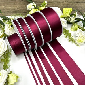 May include: A set of six burgundy satin ribbon rolls of varying widths. The rolls are arranged in a stack, with the widest roll at the bottom and the narrowest roll at the top. The ribbon is smooth and shiny, and the color is a deep, rich burgundy.