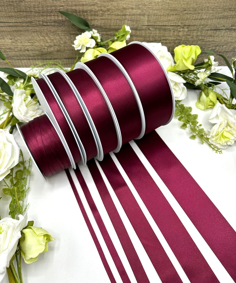 May include: A set of six burgundy satin ribbon rolls of varying widths. The rolls are arranged in a stack, with the widest roll at the bottom and the narrowest roll at the top. The ribbon is smooth and shiny, and the color is a deep, rich burgundy.