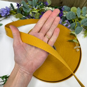 May include: Mustard yellow cotton webbing ribbon. The ribbon is tightly woven and appears to be on a roll. The ribbon is being held in a hand. The background includes greenery and purple flowers.
