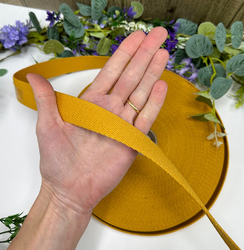 May include: Mustard yellow cotton webbing ribbon. The ribbon is tightly woven and appears to be on a roll. The ribbon is being held in a hand. The background includes greenery and purple flowers.