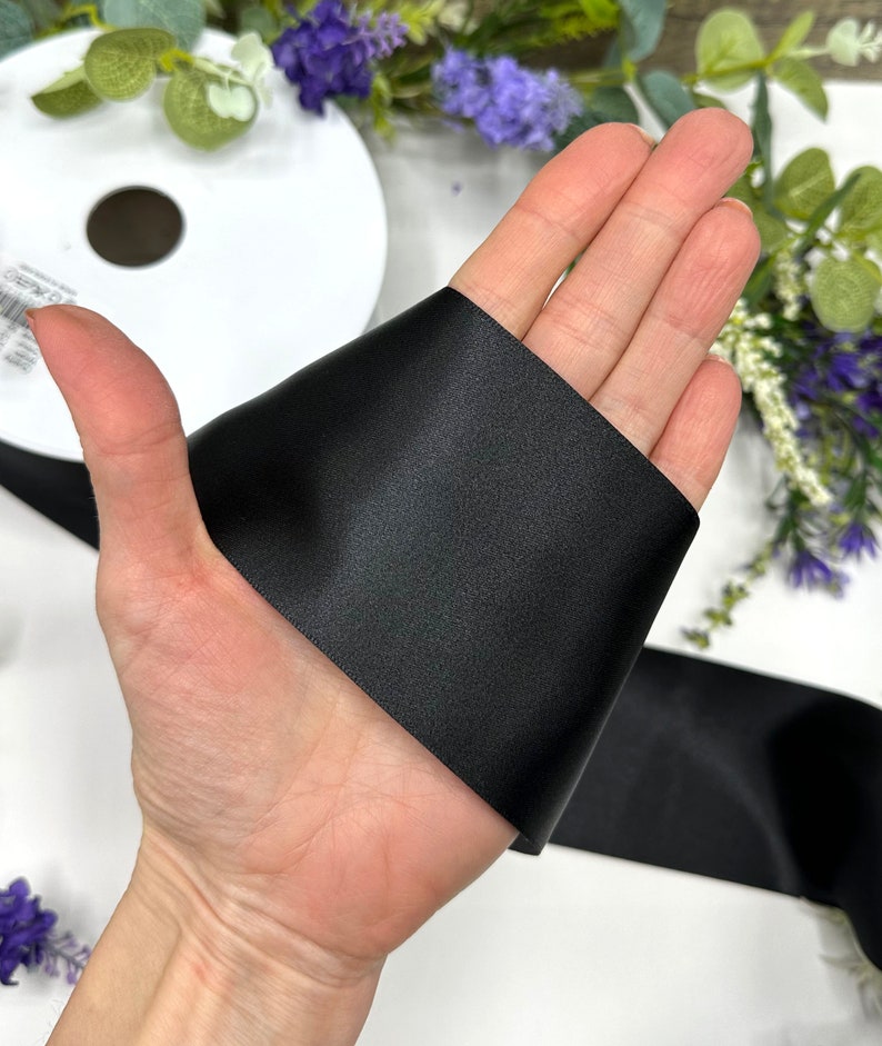 May include: A hand holding a black satin ribbon. The ribbon is folded in half and the hand is holding the folded edge.