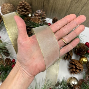 Puede incluir: Una cinta dorada con textura de malla y ribete dorado se sostiene en una mano. La cinta forma parte de una decoraci&oacute;n navide&ntilde;a, con pi&ntilde;as, bayas y adornos en el fondo. La cinta mide aproximadamente 5 cm de ancho.