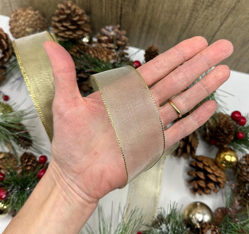 Puede incluir: Una cinta dorada con textura de malla y ribete dorado se sostiene en una mano. La cinta forma parte de una decoraci&oacute;n navide&ntilde;a, con pi&ntilde;as, bayas y adornos en el fondo. La cinta mide aproximadamente 5 cm de ancho.