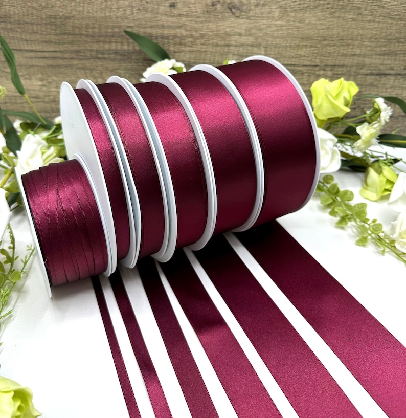 May include: A set of six spools of burgundy satin ribbon. The ribbon is arranged in a spiral, with the ends of the ribbon trailing off to the right. The ribbon is a deep, rich burgundy color.