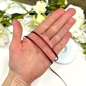 May include: Two strands of burgundy satin ribbon, approximately 1/8 inch wide, are held in a hand. The ribbon is smooth and shiny.