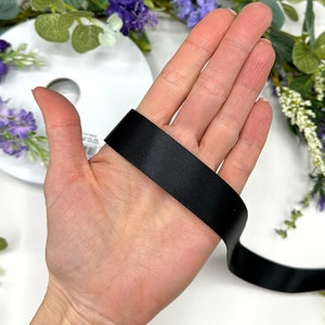 May include: A black satin ribbon is held in a hand, showing the width and texture of the ribbon.