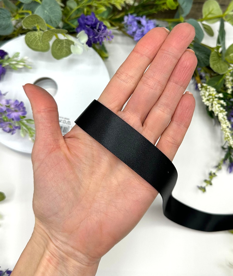 May include: A black satin ribbon is held in a hand, showing the width and texture of the ribbon.