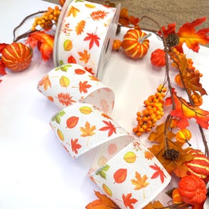 May include: A spool of white ribbon featuring a repeating pattern of autumn leaves in shades of orange, red, yellow, and green. The ribbon is displayed alongside a decorative garland of artificial orange pumpkins, brown pinecones, and fall foliage.