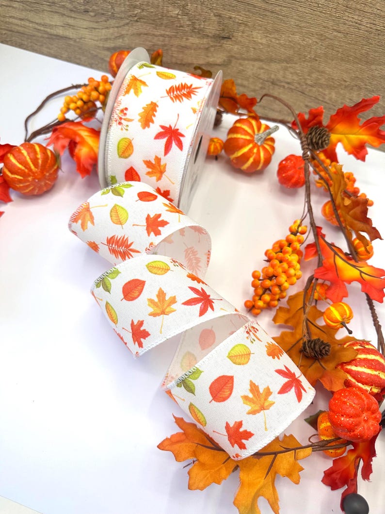 May include: A spool of white ribbon featuring a repeating pattern of autumn leaves in shades of orange, red, yellow, and green. The ribbon is displayed alongside a decorative garland of artificial orange pumpkins, brown pinecones, and fall foliage.