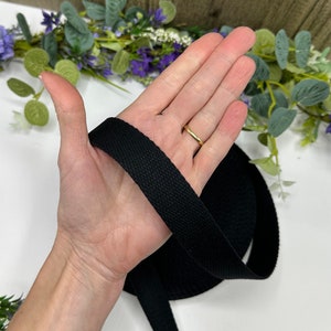 May include: A roll of black cotton webbing, held in a hand. The webbing is tightly woven and appears to be several inches wide. The background includes greenery and purple flowers.
