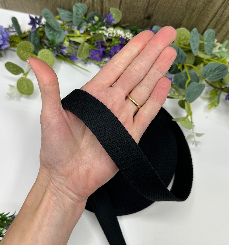 May include: A roll of black cotton webbing, held in a hand. The webbing is tightly woven and appears to be several inches wide. The background includes greenery and purple flowers.