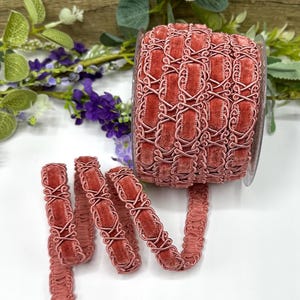 Könnte beinhalten: Eine Rolle aus altrosa Samtband mit einem aufwendigen Design. Das Band weist eine Reihe von verbundenen, länglichen Ovalen auf, die von einer zarten, helleren rosa Bordüre umrahmt werden. Das Band wird mit ein paar abgerollten Längen präsentiert, die seine Textur und Details zeigen.