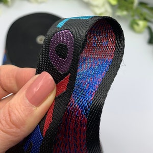 May include: Black woven webbing with red, blue, and purple accents. The webbing has a repeating pattern of a circle with a smaller circle inside. The webbing is folded in half, showing the inside of the fabric.