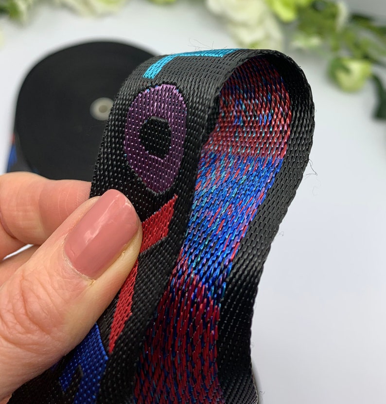 May include: Black woven webbing with red, blue, and purple accents. The webbing has a repeating pattern of a circle with a smaller circle inside. The webbing is folded in half, showing the inside of the fabric.