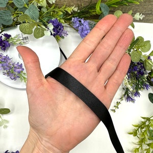 May include: A hand holding a black satin ribbon. The ribbon is about 1 inch wide and is being held in the palm of the hand.