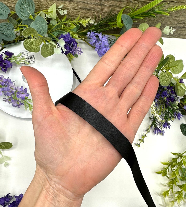 May include: A hand holding a black satin ribbon. The ribbon is about 1 inch wide and is being held in the palm of the hand.