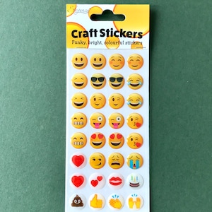 May include: A sheet of craft stickers featuring various yellow emoji faces with different expressions, including sunglasses, hearts, and tears. The packaging reads "Craft Stickers: Funky, bright, colourful stickers."