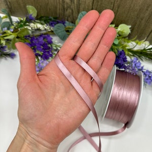 May include: A spool of dusty rose satin ribbon. The ribbon is shown wrapped around a hand and a spool.