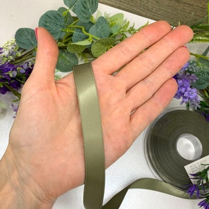 May include: A spool of olive green satin ribbon. The ribbon is approximately 1 inch wide and is shown on a hand.