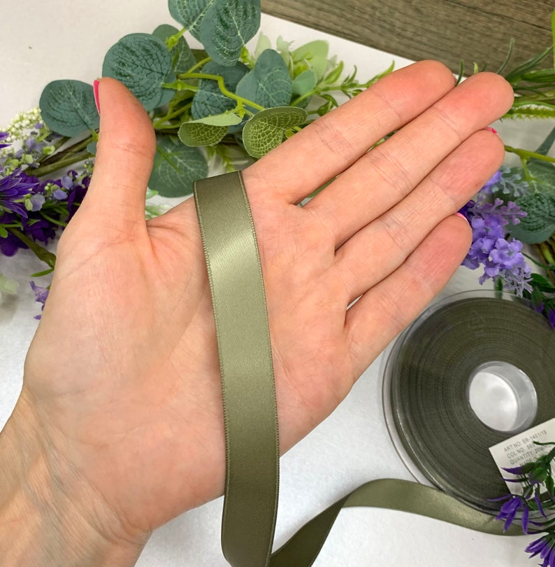 May include: A spool of olive green satin ribbon. The ribbon is approximately 1 inch wide and is shown on a hand.
