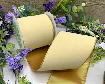Puede incluir: Un rollo de cinta de terciopelo marfil con un borde de satén dorado. La cinta está en un carrete blanco y está rodeada de flores verdes y moradas.