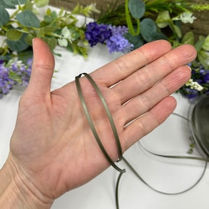 May include: A close-up of a spool of olive green satin ribbon. The ribbon is thin and has a subtle sheen.