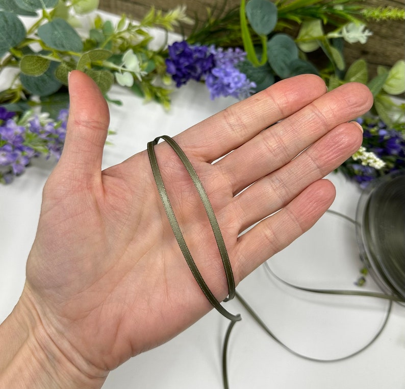 May include: A close-up of a spool of olive green satin ribbon. The ribbon is thin and has a subtle sheen.