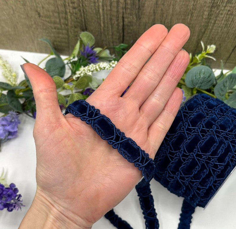 May include: A length of navy blue velvet trim with an intricate geometric design. The trim is held in a hand, showcasing its texture and detail. A roll of the trim is visible in the background, along with floral accents.
