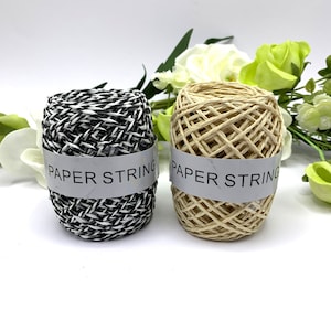 Puede incluir: Dos carretes de cuerda de papel, uno blanco y negro, el otro beige natural. Ambos carretes tienen la etiqueta "PAPER STRING" impresa.