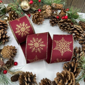 Puede incluir: Un rollo de cinta burdeos con un diseño de copo de nieve dorado y ribete dorado. La cinta se presenta entre piñas, follaje y bayas rojas, creando un ambiente festivo y de temporada.