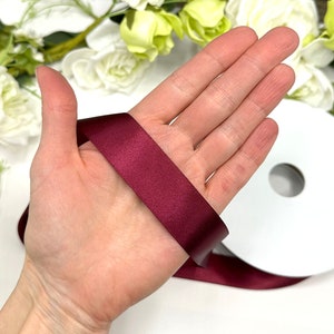 May include: A roll of burgundy satin ribbon. The ribbon is shown wrapped around a hand.