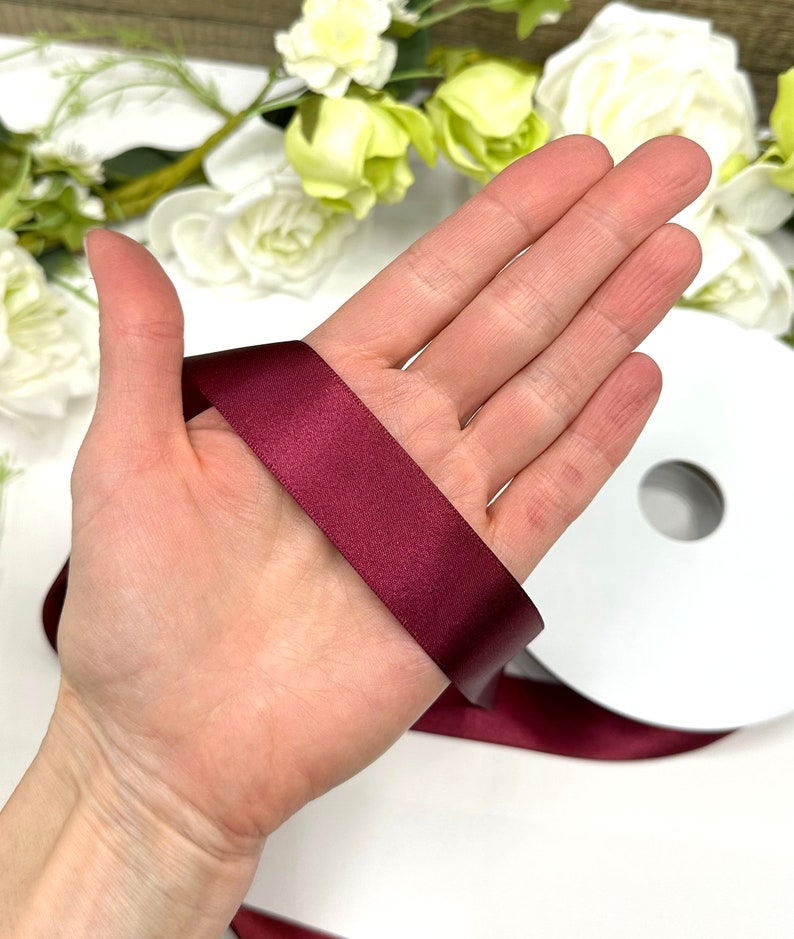 May include: A roll of burgundy satin ribbon. The ribbon is shown wrapped around a hand.