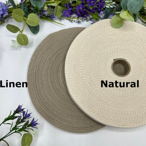 May include: Two spools of woven fabric ribbon, one in a natural off-white color and the other in a linen-colored shade of gray. The word "Natural" is printed on the right spool, and "Linen" is printed on the left spool.