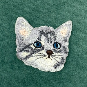 Può includere: Toppa ricamata di un gatto bianco e grigio con occhi azzurri. Il viso del gatto è ricamato in dettaglio, con un naso marrone e baffi.