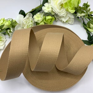 May include: A roll of beige cotton webbing, a type of strong fabric tape, is shown on a white surface. The webbing is approximately 1 inch wide and has a textured surface.