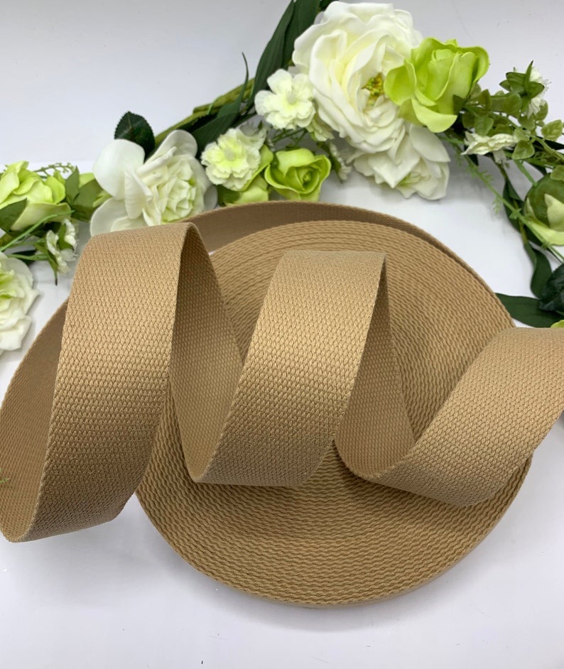 May include: A roll of beige cotton webbing, a type of strong fabric tape, is shown on a white surface. The webbing is approximately 1 inch wide and has a textured surface.